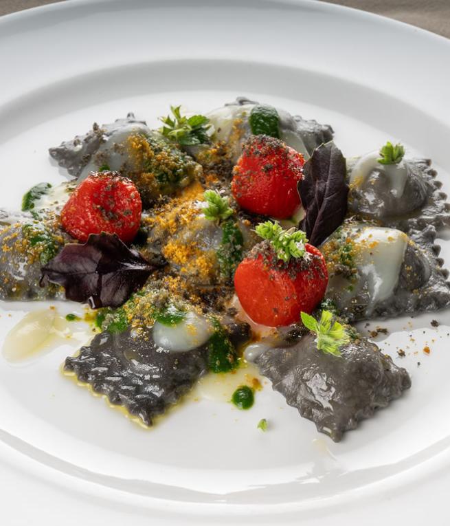 Ravioli neri con pomodorini rossi, erbe fresche e salse colorate, un'esplosione di sapori.